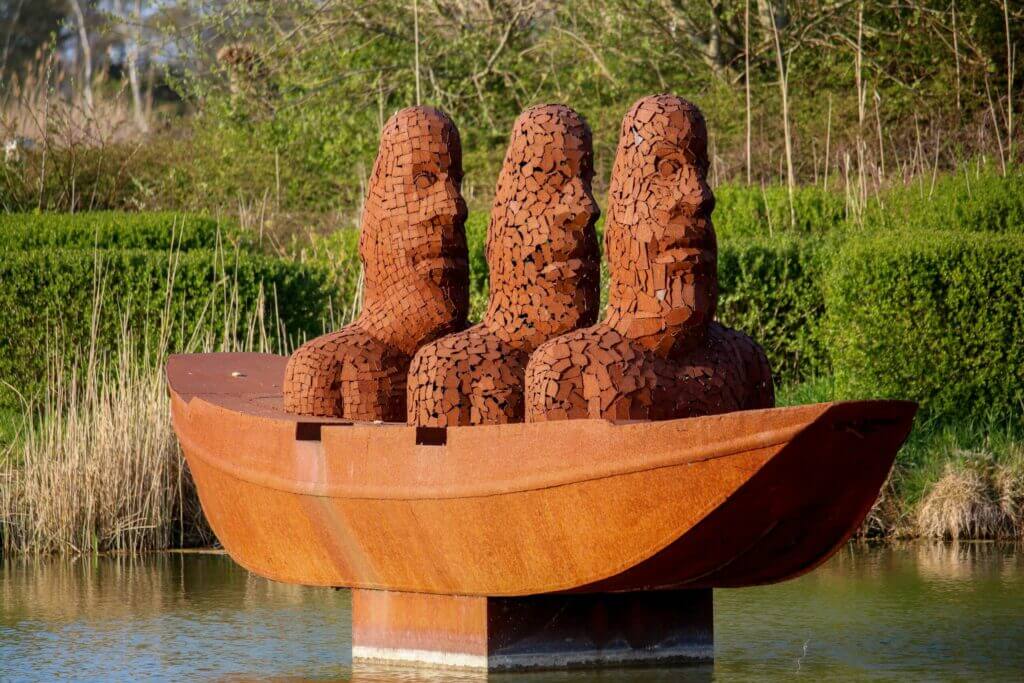 Artistic sculpture of three figures in a rusted boat located in Lommel, Belgium, surrounded by nature.